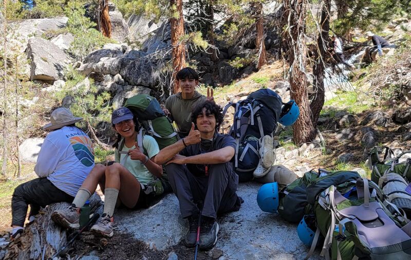 The image shows a group of young hikers resting outdoors. They are surrounded by backpacks and appear to be taking a break on a rocky terrain, possibly during a hiking trip. Some of them are giving a thumbs-up, suggesting a positive and energetic atmosphere. The background features trees and natural scenery, indicating they are in a forest or mountainous area.